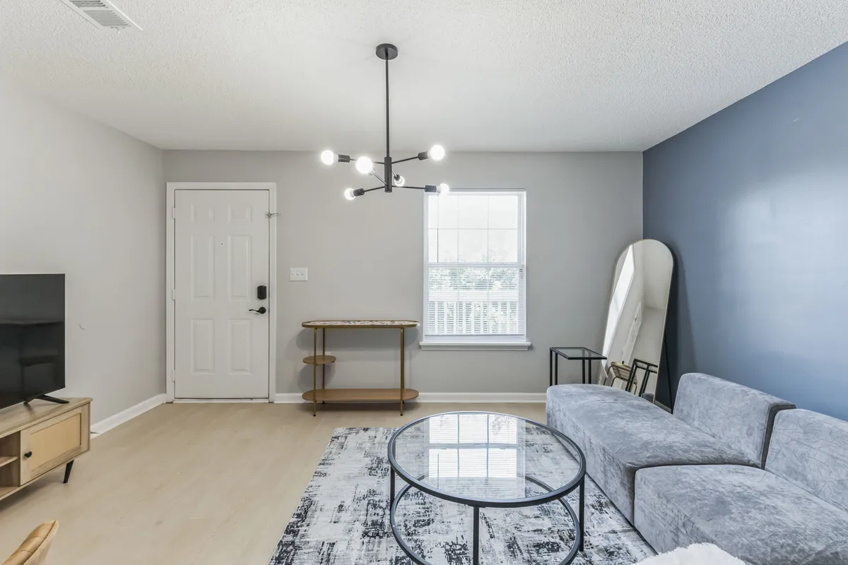A welcoming living area featuring a modern light fixture, a comfortable sofa, a coffee table, a decorative wall mirror, and a bright entry door. The setting is perfect for both quiet evenings and intimate gatherings, with soft lighting and tasteful decor enhancing the inviting mood.