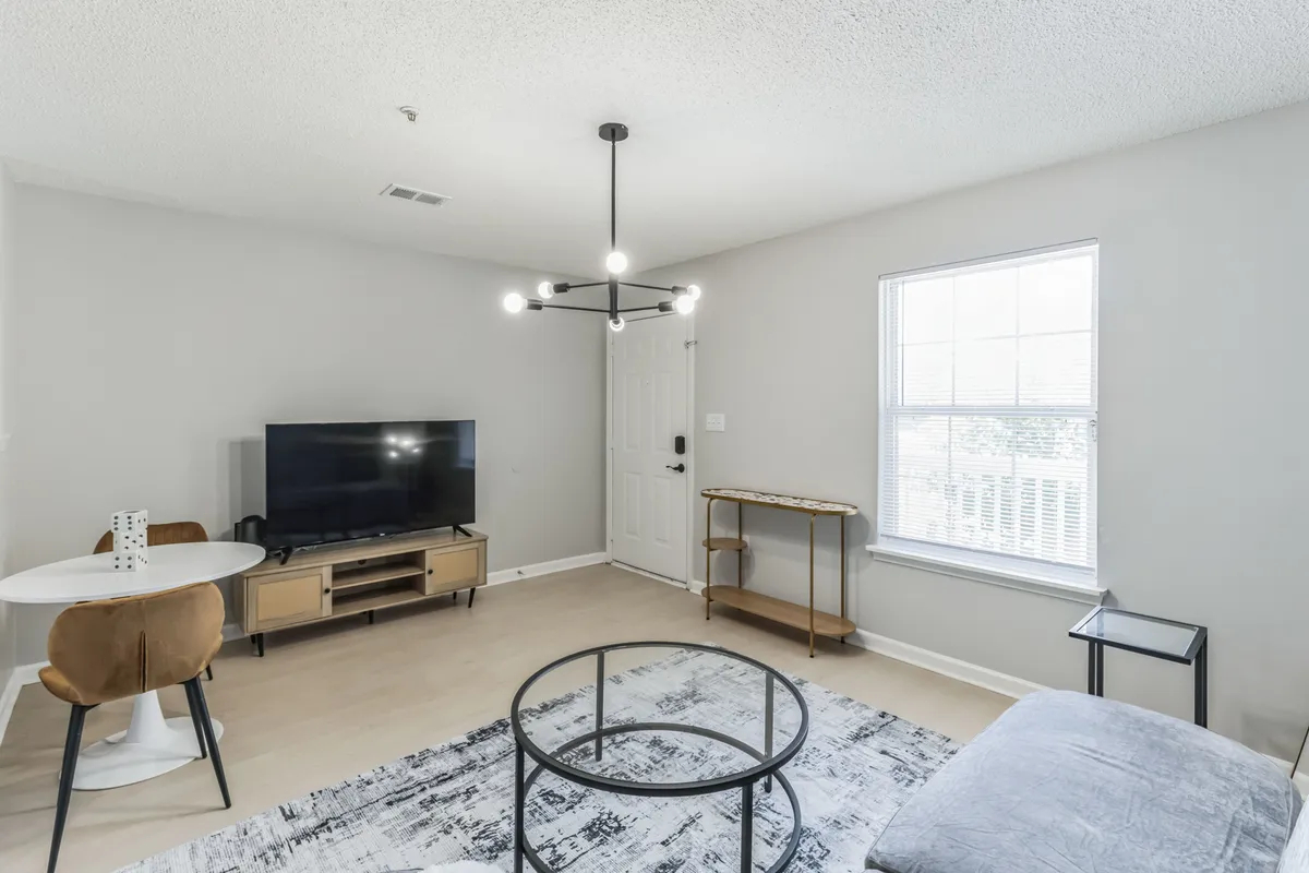 A soft, comfortable living space with a large sofa, circular coffee table, television, dining table, and decorative lighting. The arrangement is open and inviting, illuminated by natural light and designed for both relaxation and casual socializing.