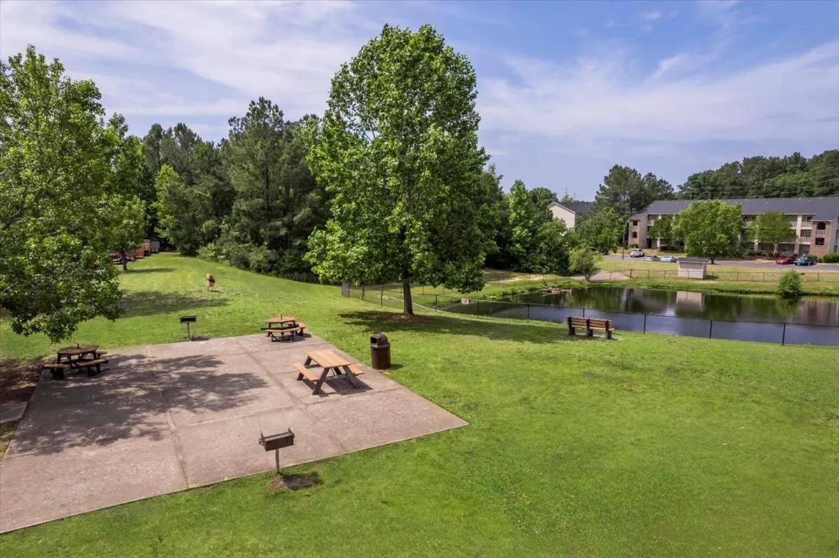 A tranquil exterior scene featuring a lush pond surrounded by trees and green grass. Benches are scattered around, and people are seen enjoying picnics, walking along the path, fishing by the water, and children playing on the grass. The atmosphere is peaceful, with a hint of early morning or sunset light reflecting on the water.