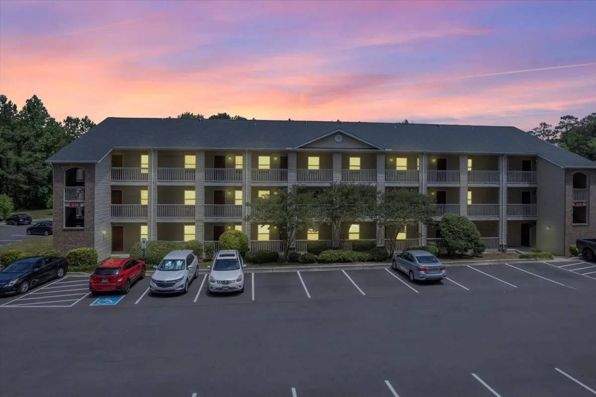 The image shows the apartment building at sunset, with a soft glow highlighting the structure, parking lot, and mature trees. Residents can be seen gathering and enjoying the evening outside, suggesting a relaxed, home-like environment.