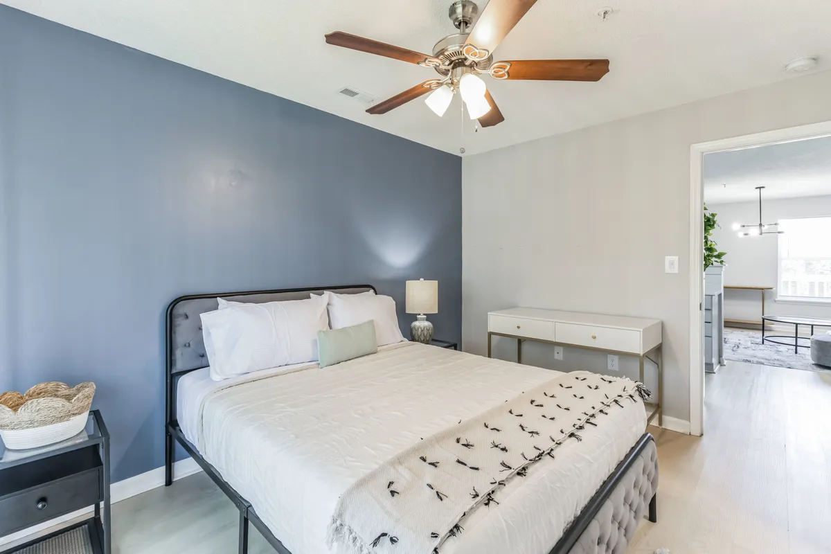 A tranquil bedroom scene with a cozy queen bed, soft lighting from a bedside lamp, and a ceiling fan overhead. The modern nightstand and uncluttered design evoke a restful, hotel-quality environment perfect for unwinding after a busy day.