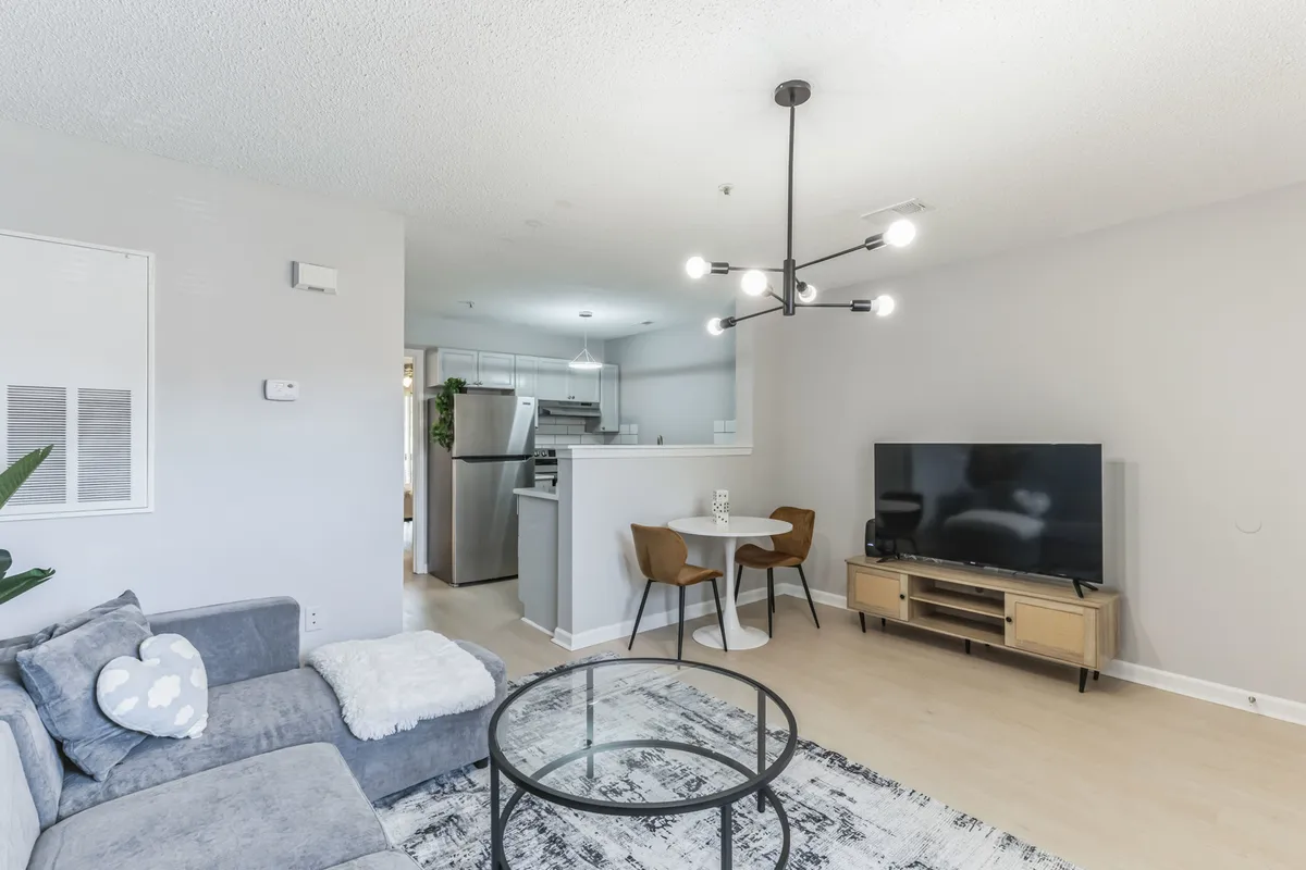 A cozy and contemporary living area featuring a plush sofa adorned with decorative pillows, a round glass coffee table, a modern light fixture overhead, a streaming-ready television, and a view into the adjoining dining area with stylish chairs. The room is bathed in natural light from large windows, creating an airy and inviting atmosphere.