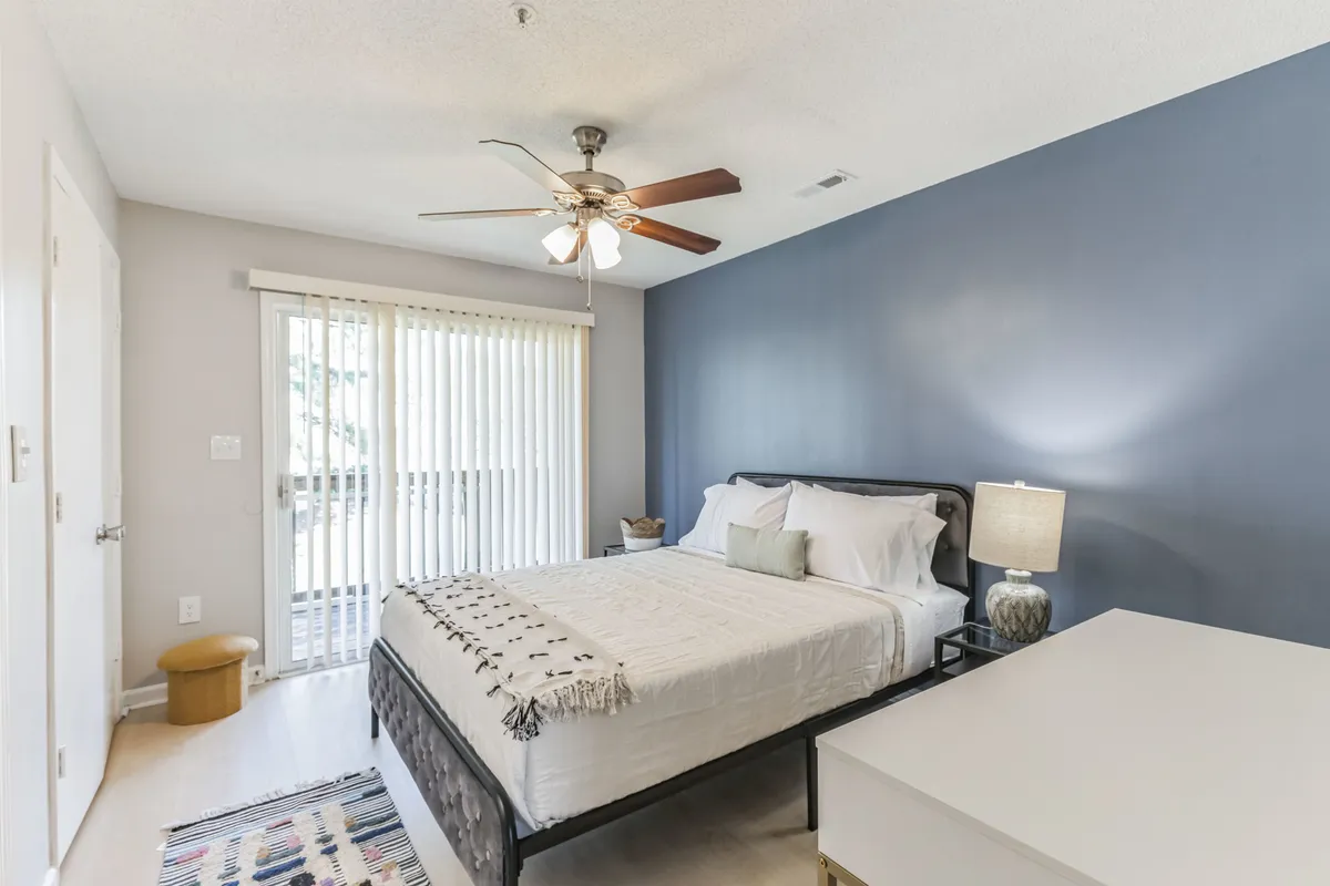 A bright, serene bedroom featuring a plush queen bed dressed in inviting linens. A ceiling fan hangs above for added comfort, while natural light filters through window blinds. The cozy nightstand with a lamp invites bedside reading or unwinding after a long day, and the overall modern décor sets a calming atmosphere.