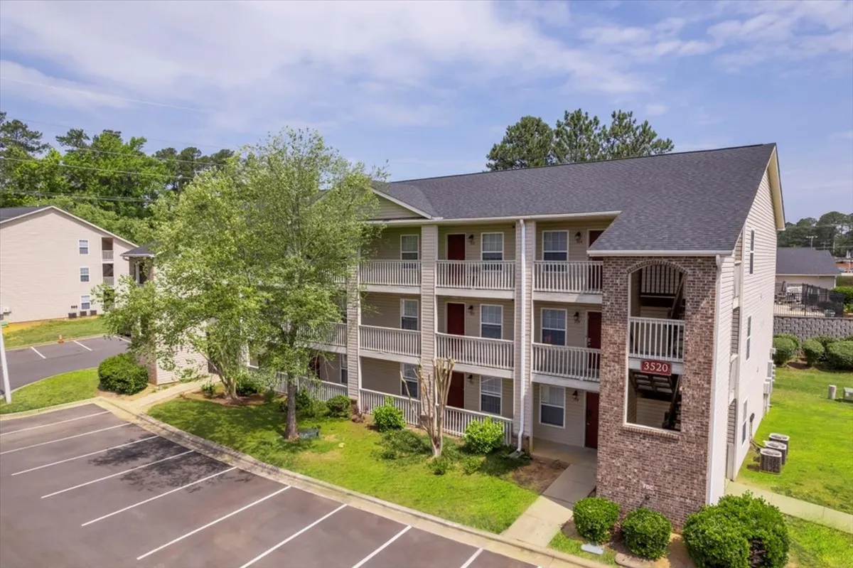 A three-story apartment building is surrounded by mature green trees with a neatly maintained parking lot in the foreground. The setting feels inviting, with residents visible outdoors and a sense of community in the landscaped green spaces.