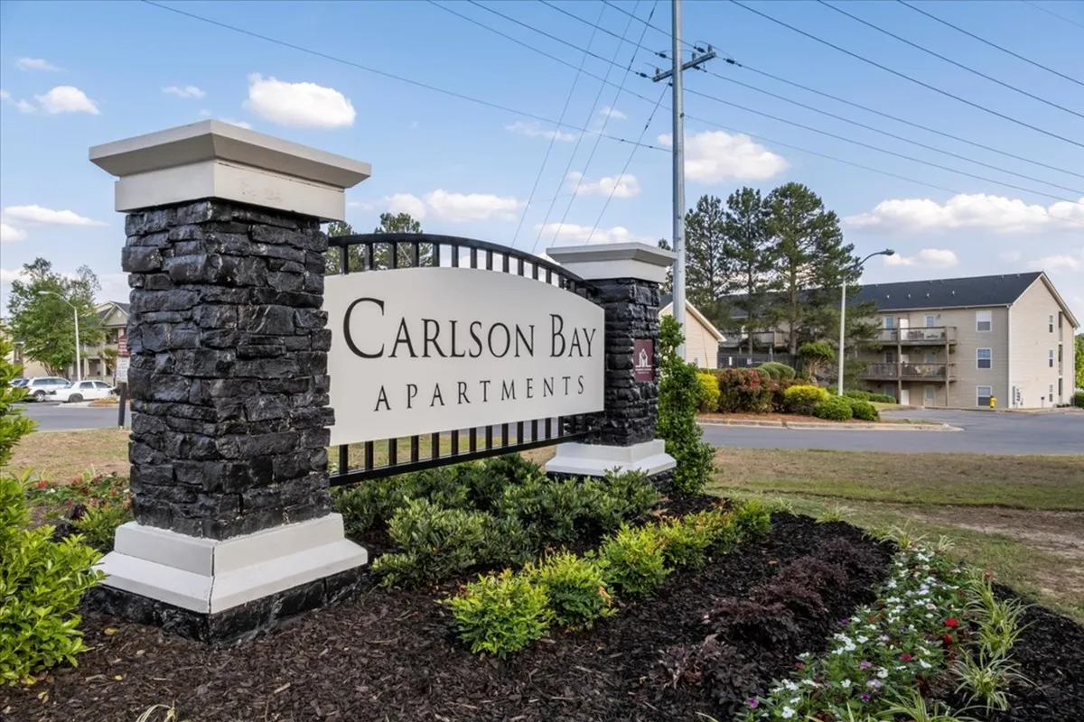 The image displays the front of the Carlson Bay apartment community, featuring inviting signage, attractive landscaping, and the building’s welcoming structure. People are seen enjoying the outdoor areas, suggesting a friendly and accessible environment.