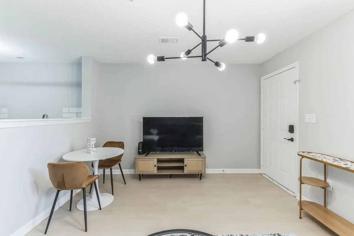 A multifunctional dining area with a modern dining table beneath a chandelier, a television for entertainment, and an entry door nearby. The room’s layout encourages socializing, shared meals, and relaxed evenings at home.